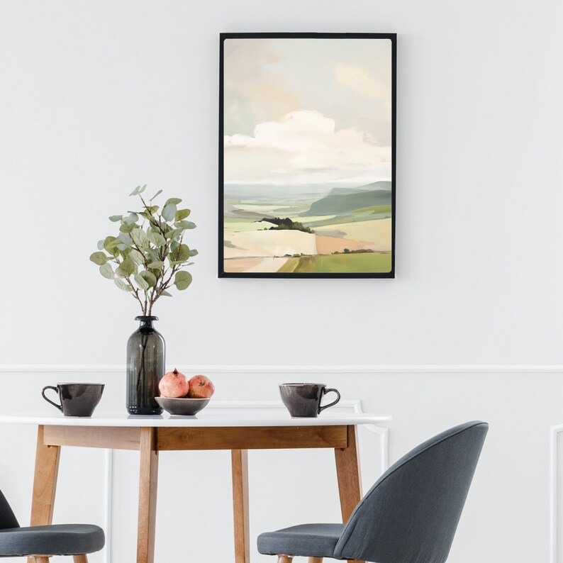 Puede incluir: Una pintura de paisaje enmarcada con un cielo azul claro y nubes blancas sobre un campo verde y marr&oacute;n. La pintura est&aacute; colgada en una pared blanca sobre una mesa de madera con dos tazas y un cuenco de fruta.