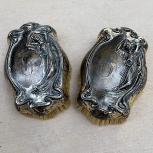May include: Two antique silver-toned hair brushes. Each brush features an oval design with a detailed relief of a woman and ornate scrollwork. The bristles are a light tan color. The brushes are likely from the early 20th century.