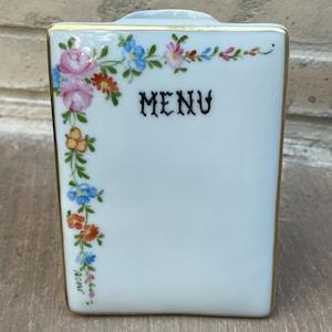 May include: A white porcelain menu holder with a gold trim. The front features the word "MENU" in black script and is decorated with a floral design in pink, blue, orange, and yellow. The holder has a rectangular shape.