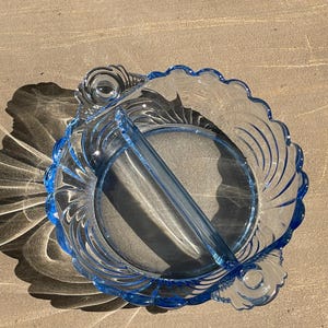 May include: A clear, divided glass serving dish with a light blue trim. The dish has a scalloped edge and a central divider. The design includes curved lines and decorative accents, creating an elegant look.