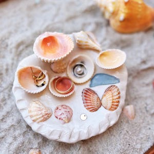 May include: A white ceramic ring dish adorned with various seashells, including pink, orange, and cream-colored varieties. The dish holds gold rings and a small earring. The arrangement is set on a bed of sand, evoking a beach theme.