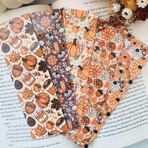 May include: A collection of fall-themed bookmarks with various patterns. Designs include pumpkins, flowers, acorns, and leaves in warm orange, brown, and cream colors. The bookmarks are arranged on an open book, suggesting a literary theme.