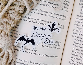 May include: A black and white sticker with a dragon and the text "In my Dragon Era" on a white background. The sticker is placed on a book with text about a person walking to a bedroom door.