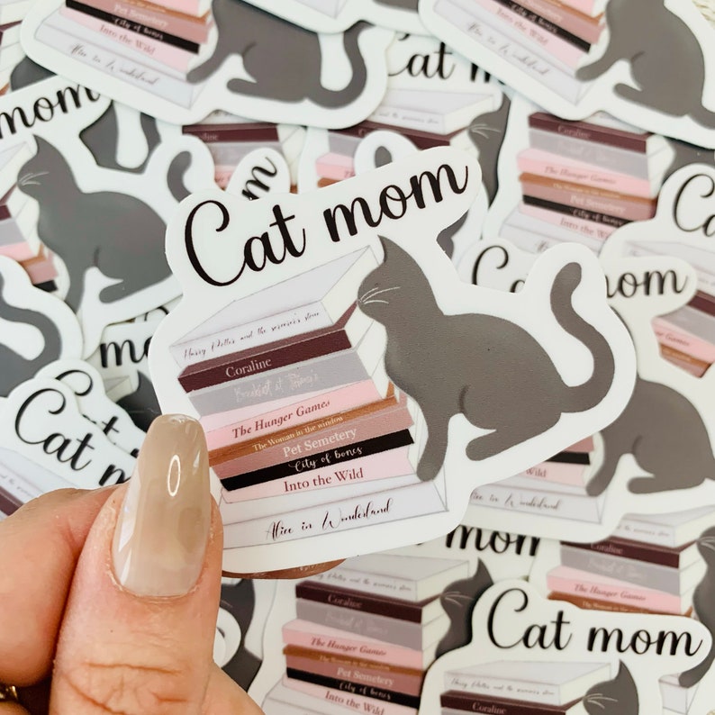 Op de afbeelding: Een sticker met een grijze kat die op een stapel boeken zit. De boeken hebben titels zoals "Coraline", "The Hunger Games", "Pet Semetery", "City of Bones", "Into the Wild" en "Alice in Wonderland". De sticker heeft de tekst "Cat Mom" in zwarte letters.