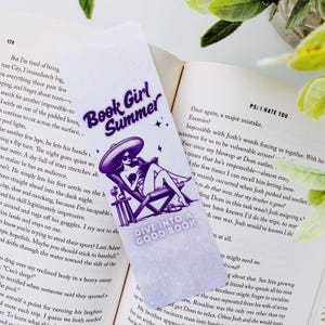 以下が含まれることがあります： 「Book Girl Summer」の文字と、ビーチチェアに座る女性のイラストが描かれた紫と白のブックマーク。ブックマークには「Dive into a Good Book」の文字も。ブックマークは開いた本の上に置かれています。