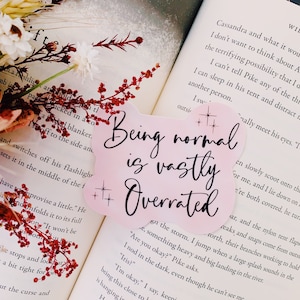 May include: Pink and white motivational quote sticker with the text "Being normal is vastly overrated" in black script font. The sticker has two small gold stars on either side of the text.