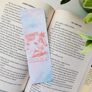 以下が含まれることがあります： 水彩効果のあるブックマーク。テキスト「SUMMER SMUT」と、ビーチで読書をする女性のイラストが特徴です。デザインにはヤシの木と「KISSED BY THE SUN and spanked by the plot」というフレーズが含まれています。