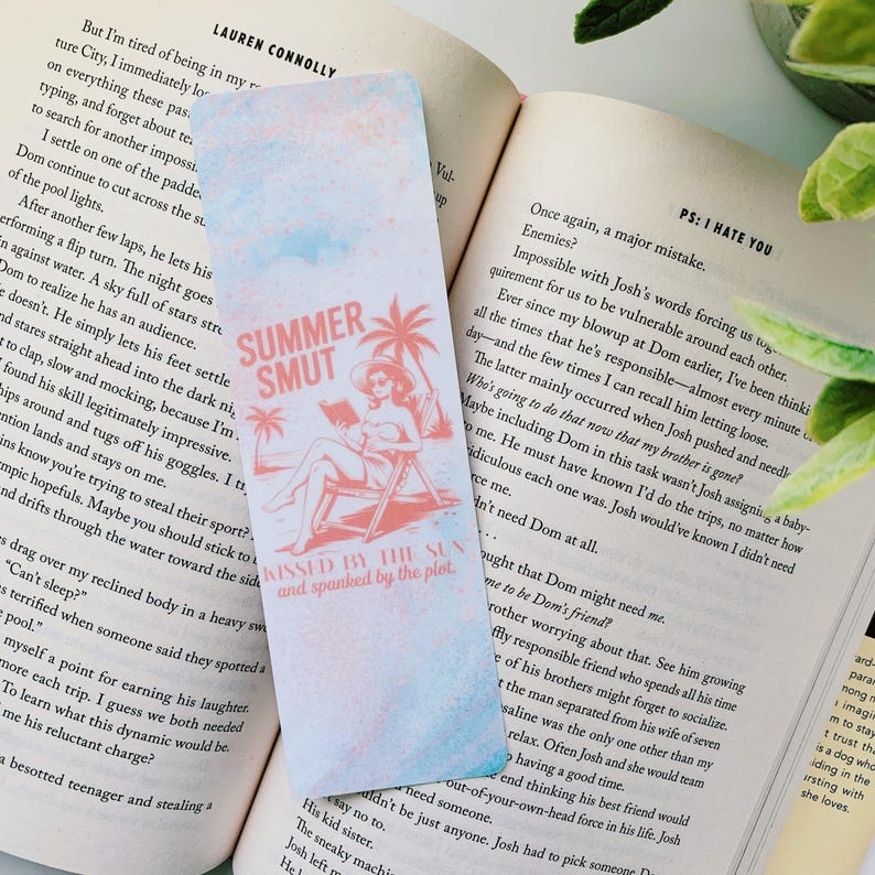 以下が含まれることがあります： 水彩効果のあるブックマーク。テキスト「SUMMER SMUT」と、ビーチで読書をする女性のイラストが特徴です。デザインにはヤシの木と「KISSED BY THE SUN and spanked by the plot」というフレーズが含まれています。