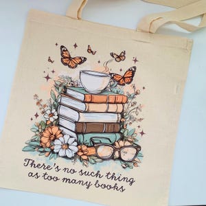 May include: A beige tote bag with a whimsical illustration of a stack of books, a cup of coffee, butterflies, and flowers. The text "There's no such thing as too many books" is printed on the bag.