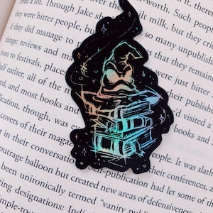 May include: Black holographic sticker featuring a witch hat and a stack of books. The design includes stars and a subtle rainbow gradient. The sticker is placed on an open book with printed text.