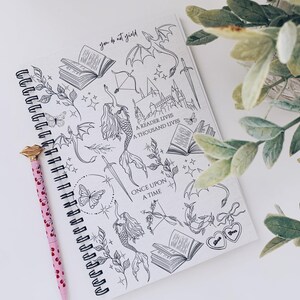 May include: Spiral-bound notebook with black and white fantasy illustrations, including mermaids, dragons, and books. A pink pen with a gold-colored tip rests beside the notebook. Text on the notebook reads "you do not yield" and "A READER LIVES A THOUSAND LIVES".