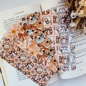 May include: A collection of decorative bookmarks with fall-themed patterns. Designs include pumpkins, bows, and floral arrangements in shades of orange, peach, and white. The bookmarks are displayed open on a book.