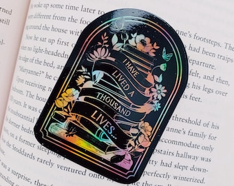 May include: A black holographic sticker with a stack of books, flowers, and a banner that reads "I have lived a thousand lives..." The sticker has a rainbow iridescent finish.