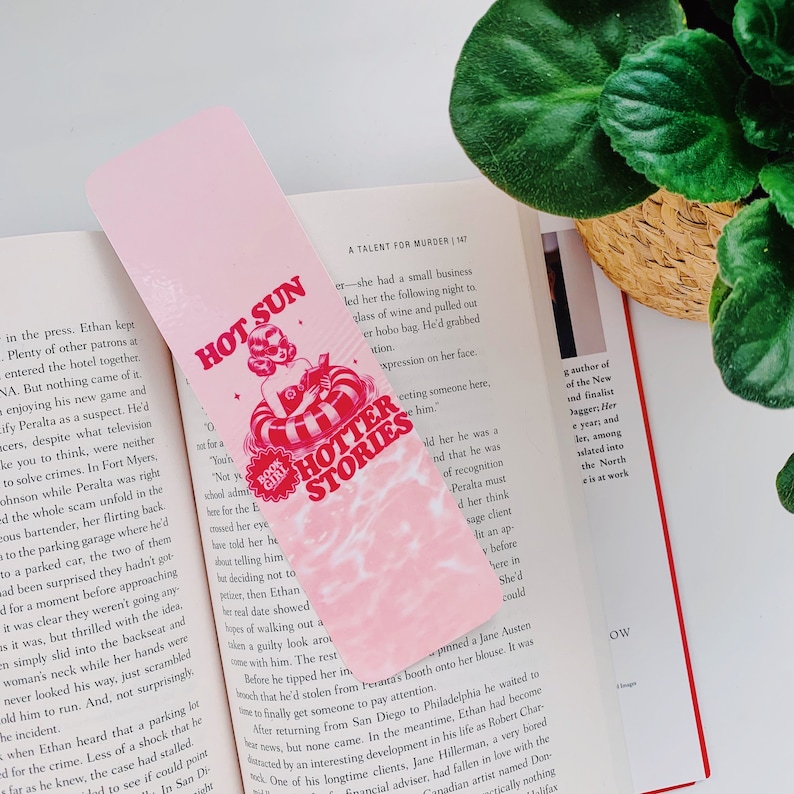 May include: Pink bookmark with the text "HOT SUN HOTTER STORIES" and an illustration of a woman in a pool float. The bookmark is placed in an open book.