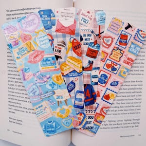 Puede incluir: Un conjunto de marcap&aacute;ginas coloridos y decorativos con varios dise&ntilde;os y texto. Los marcap&aacute;ginas presentan ilustraciones de corazones, alimentos y frases como "See You Next Season" e "I Need You". Se encuentran dentro de un libro abierto.