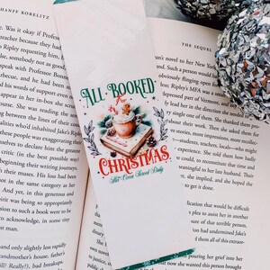 May include: A Christmas-themed bookmark with the text "ALL BOOKED for CHRISTMAS". The bookmark features a festive design with a cup of hot cocoa, gingerbread man, and pine boughs. It is placed in an open book.