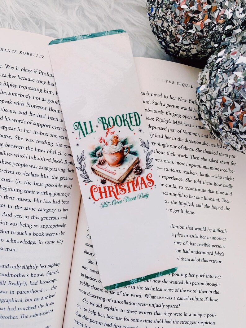 May include: A Christmas-themed bookmark with the text "ALL BOOKED for CHRISTMAS". The bookmark features a festive design with a cup of hot cocoa, gingerbread man, and pine boughs. It is placed in an open book.