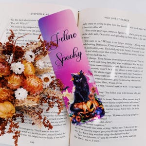 K&ouml;nnte beinhalten: Ein lila und pinkfarbenes Lesezeichen mit einer schwarzen Katze, die auf einem K&uuml;rbis sitzt. Das Lesezeichen tr&auml;gt die Aufschrift "Feline Spooky". Der Text "You Like It Darker" ist im Hintergrund zu sehen.