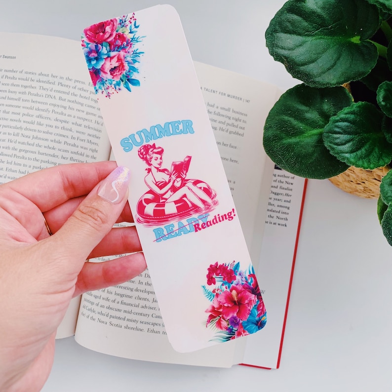 May include: A decorative bookmark with a floral design and the text "Summer Read Reading!". The bookmark features a retro illustration of a woman in a pool float reading a book. The design includes pink, blue, and white colors.