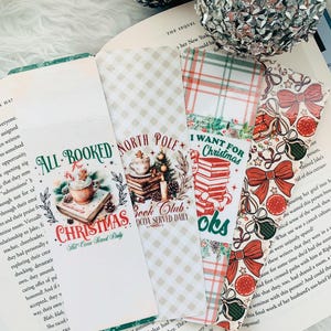 May include: A collection of Christmas-themed bookmarks with various designs. One bookmark reads "All Booked Christmas Hot Cocoa Served Daily" with a cocoa cup graphic. Other bookmarks feature plaid, bows, and Christmas ornaments. The bookmarks are placed on an open book.