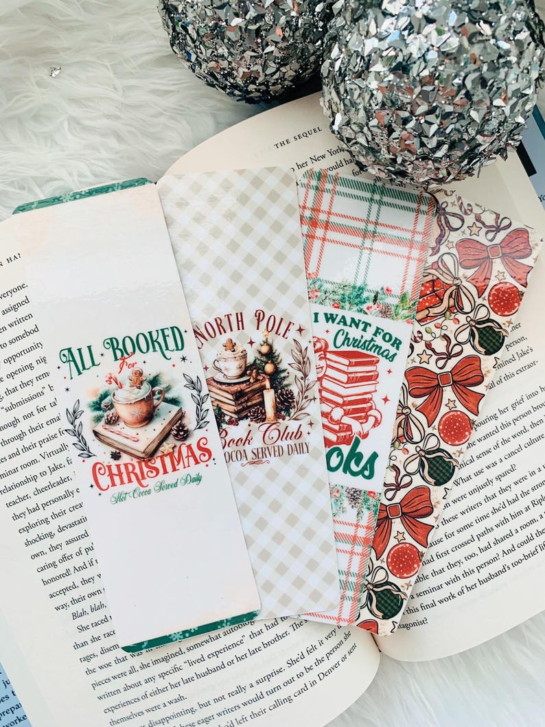 May include: A collection of Christmas-themed bookmarks with various designs. One bookmark reads "All Booked Christmas Hot Cocoa Served Daily" with a cocoa cup graphic. Other bookmarks feature plaid, bows, and Christmas ornaments. The bookmarks are placed on an open book.