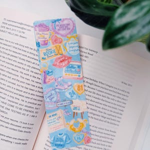 Puede incluir: Un marcap&aacute;ginas colorido con varias ilustraciones y texto, incluyendo corazones, una camiseta de Boston y las palabras "I need you". El marcap&aacute;ginas est&aacute; colocado en un libro abierto.