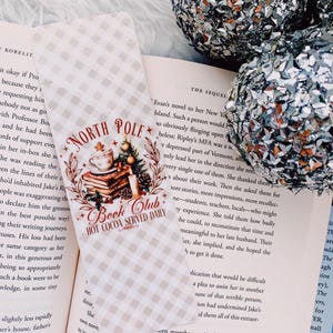 May include: A bookmark with the text "North Pole Book Club Hot Cocoa Served Daily" over a stack of books and a Christmas tree. The bookmark is on a book with a checkered pattern. Two silver glitter ornaments are in the background.