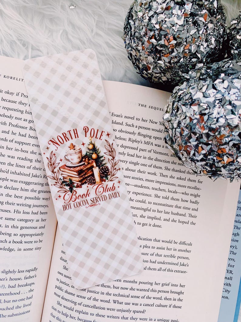 May include: A bookmark with the text "North Pole Book Club Hot Cocoa Served Daily" over a stack of books and a Christmas tree. The bookmark is on a book with a checkered pattern. Two silver glitter ornaments are in the background.