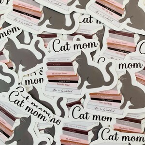Op de afbeelding: Een verzameling stickers met een grijze kat die op een stapel boeken zit. De boeken hebben titels zoals "The Hunger Games", "Pet Semetery", "City of Bones" en "Into the Wild". De stickers hebben ook de tekst "Cat Mom" erop.
