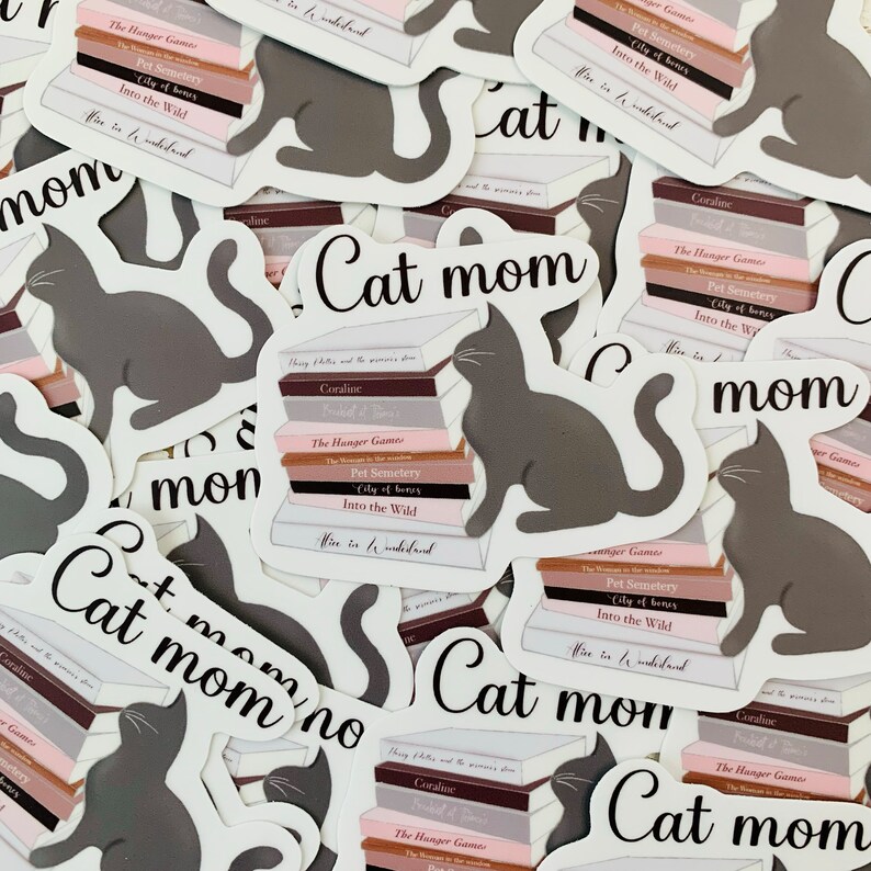 Op de afbeelding: Een verzameling stickers met een grijze kat die op een stapel boeken zit. De boeken hebben titels zoals "The Hunger Games", "Pet Semetery", "City of Bones" en "Into the Wild". De stickers hebben ook de tekst "Cat Mom" erop.