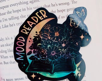 May include: A black and white sticker with a holographic finish. The sticker features a book with flowers and a crescent moon. The text "Mood Reader" is written in a curved font around the moon.