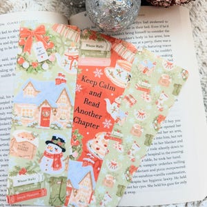 May include: Three rectangular paper bookmarks featuring festive holiday illustrations. The designs include a snowman, a snowy cottage, stacks of books, and tea sets. The center bookmark displays the text, Keep Calm and Read Another Chapter.