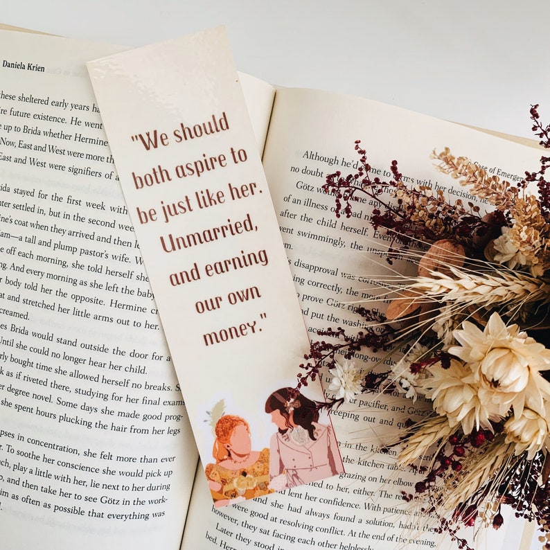 以下が含まれることがあります： ダニエラ・クリーンの著書「The Unmarried」からの引用が書かれたしおり。引用文は「私たちは彼女のようにありたいと願うべきです。結婚せず、自分のお金を稼いで。」しおりには、ヴィンテージスタイルで描かれた2人の女性のイラストが描かれています。