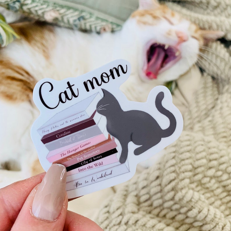 Op de afbeelding: Een witte sticker met de tekst "Cat mom" en een zwart silhouet van een kat die op een stapel boeken zit. De boektitels zijn "Harry Potter en de Steen der Wijzen", "Coraline", "Freaky & Davis", "The Hunger Games", "The Haunting in the Shadow", "Pet Semetery", "City of Bones", "Into the Wild", en "Wise in the Wilderness".