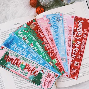 May include: A collection of colorful holiday-themed bookmarks with text like "North Pole Book Club" and "Santa's Ho Ho Ho Room." The bookmarks are decorated with festive imagery, including trees, gingerbread men, and snowflakes. Red and silver ornaments are in the background.