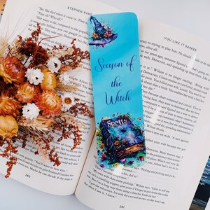 K&ouml;nnte beinhalten: Ein blaues und gr&uuml;nes Lesezeichen mit einem Hexenhut und einem Buch mit dem Titel "Spells" auf dem Cover. Das Lesezeichen tr&auml;gt den Text "Season of the Witch".