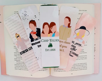 May include: Four colorful bookmarks with illustrations of characters from popular movies and TV shows. The bookmarks feature a green pine tree with the text "Camp Walden Est. 1998", a group of two women, a man holding a deck of cards, and a woman wearing a hat. The text "Whatever Crew" is visible on the bookmark with the man holding cards. The text "We have class and you don't." is visible on the bookmark with the two women. The text "VG young and atifal a CRIME Snow" is visible on the bookmark with the woman wearing a hat.