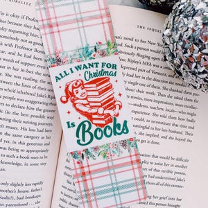 May include: A Christmas-themed bookmark with the text "All I Want For Christmas Is Books." The bookmark features a red and green plaid pattern and a graphic of a person holding a stack of books. The bookmark is placed on an open book.