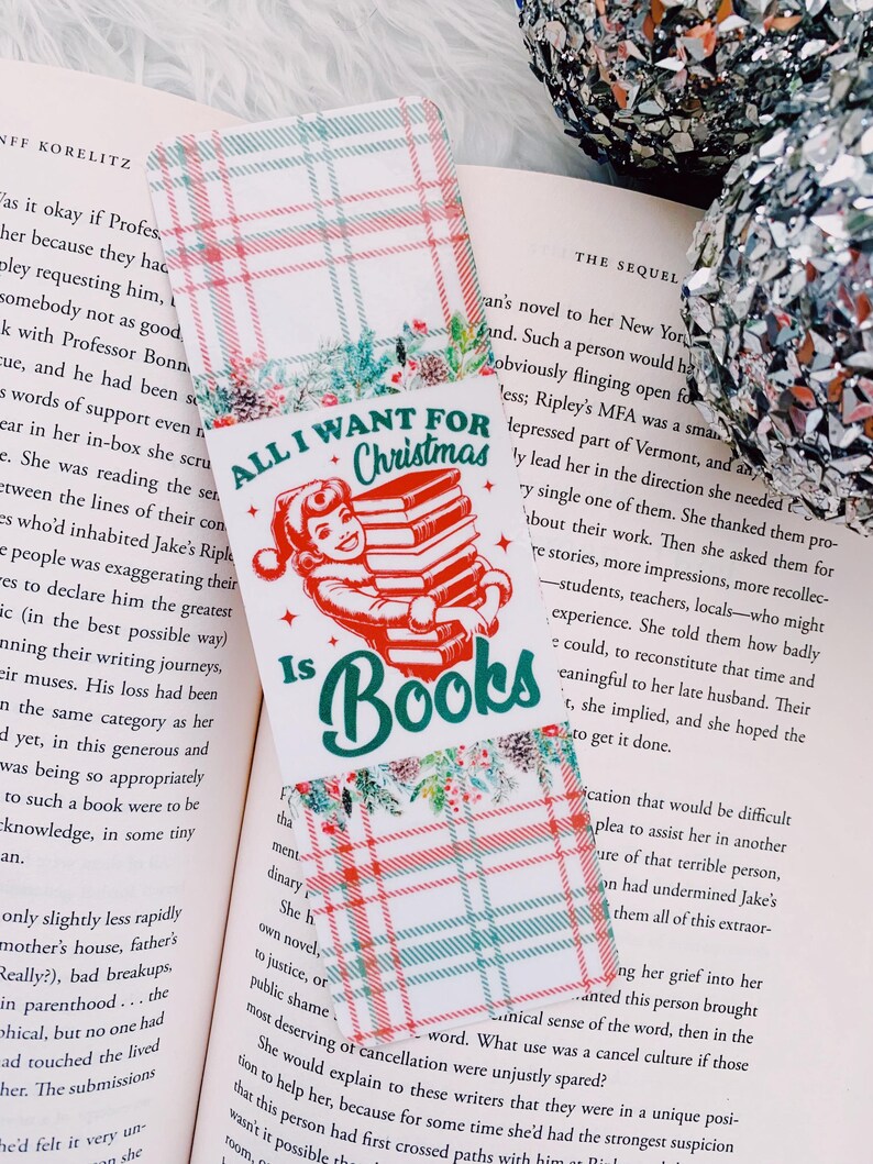 May include: A Christmas-themed bookmark with the text "All I Want For Christmas Is Books." The bookmark features a red and green plaid pattern and a graphic of a person holding a stack of books. The bookmark is placed on an open book.