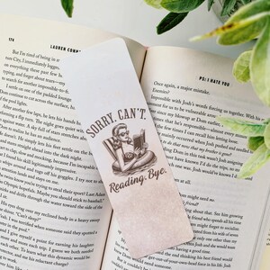 以下が含まれることがあります： サングラスをかけた女性が浮き輪に乗って本を読んでいるヴィンテージイラストの長方形のブックマーク。ブックマークのテキストは「Sorry, Can't. Reading. Bye.」です。ブックマークは開いた本の中にあります。