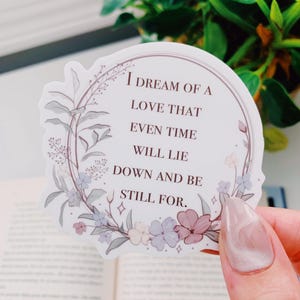 May include: A sticker with the text "I DREAM OF A LOVE THAT EVEN TIME WILL LIE DOWN AND BE STILL FOR." The sticker has a floral wreath design with pastel colors. The sticker is held in a hand.