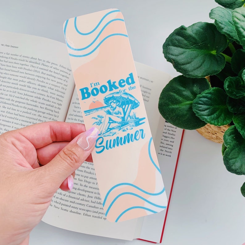 May include: A bookmark with a peach background and blue wave design. The text "I'm Booked for the Summer" is displayed with a beach scene illustration. The bookmark is held over an open book.