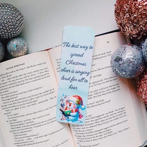 May include: A blue and white bookmark with a snowman illustration. The snowman is wearing a red Santa hat and holding a microphone. The text on the bookmark reads "The best way to spread Christmas cheer is singing loud for all to hear."