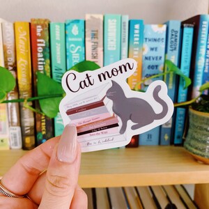 Op de afbeelding: Een sticker van een grijze kat die een stapel boeken leest. De boeken hebben titels zoals "Coraline", "The Hunger Games", "Pet Sematary", "City of Bones", "Into the Wild", en "Where the Wild Things Are". De sticker heeft bovenaan de tekst "Cat mom".