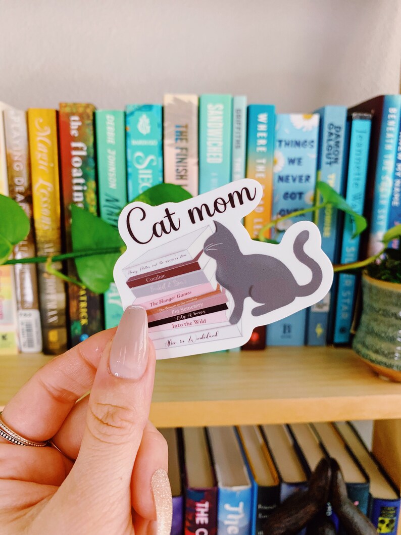 Op de afbeelding: Een sticker van een grijze kat die een stapel boeken leest. De boeken hebben titels zoals "Coraline", "The Hunger Games", "Pet Sematary", "City of Bones", "Into the Wild", en "Where the Wild Things Are". De sticker heeft bovenaan de tekst "Cat mom".