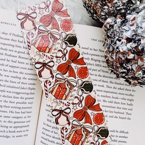 May include: A festive Christmas-themed bookmark with a white background, adorned with red bows, gift boxes, ornaments, and candy canes. The bookmark is placed on an open book, adding a touch of holiday cheer to reading.