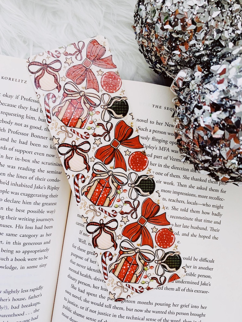 May include: A festive Christmas-themed bookmark with a white background, adorned with red bows, gift boxes, ornaments, and candy canes. The bookmark is placed on an open book, adding a touch of holiday cheer to reading.