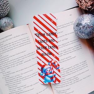 May include: A red and white striped bookmark with a snowman holding a candy cane. The bookmark has the text "With Candy Canes and Silver Lanes Aglow".