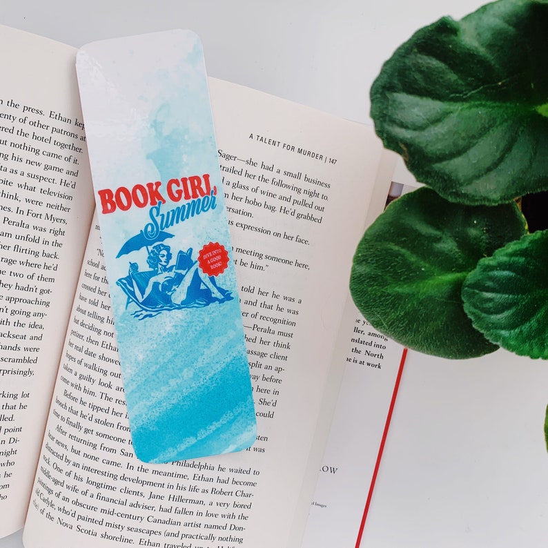 May include: A bookmark with a watercolor design featuring the text "BOOK GIRL Summer" in red and blue. The bookmark has an illustration of a person reading under an umbrella. A book and a plant are in the background.