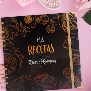 Puede incluir: Un libro de recetas de espiral negro con detalles dorados y un marcapáginas de cinta dorada. La portada presenta una ilustración dorada de varios artículos de comida y el texto "Mis Recetas Elena Rodriguez".